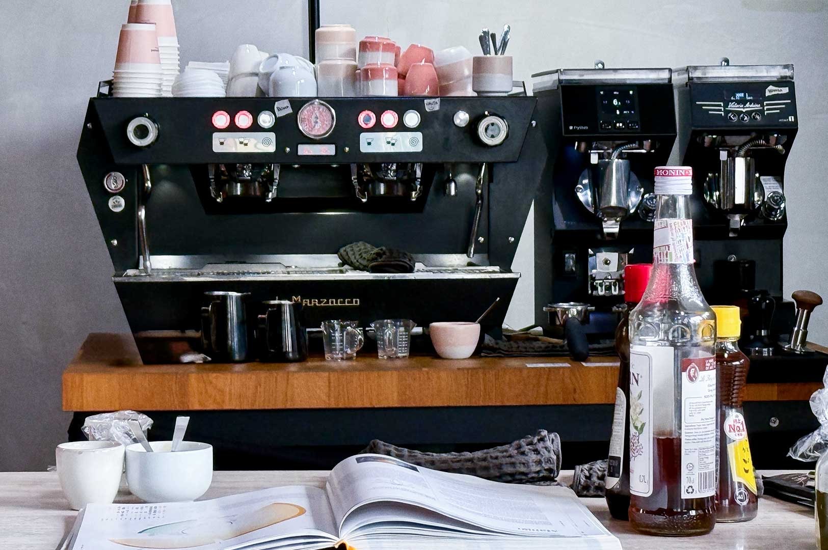 Barista workstation with a multi‑group espresso machine, pink and white cups on top, grinders labeled Fryskin and Mazzer Luigi, and pitchers and condiments arranged nearby.