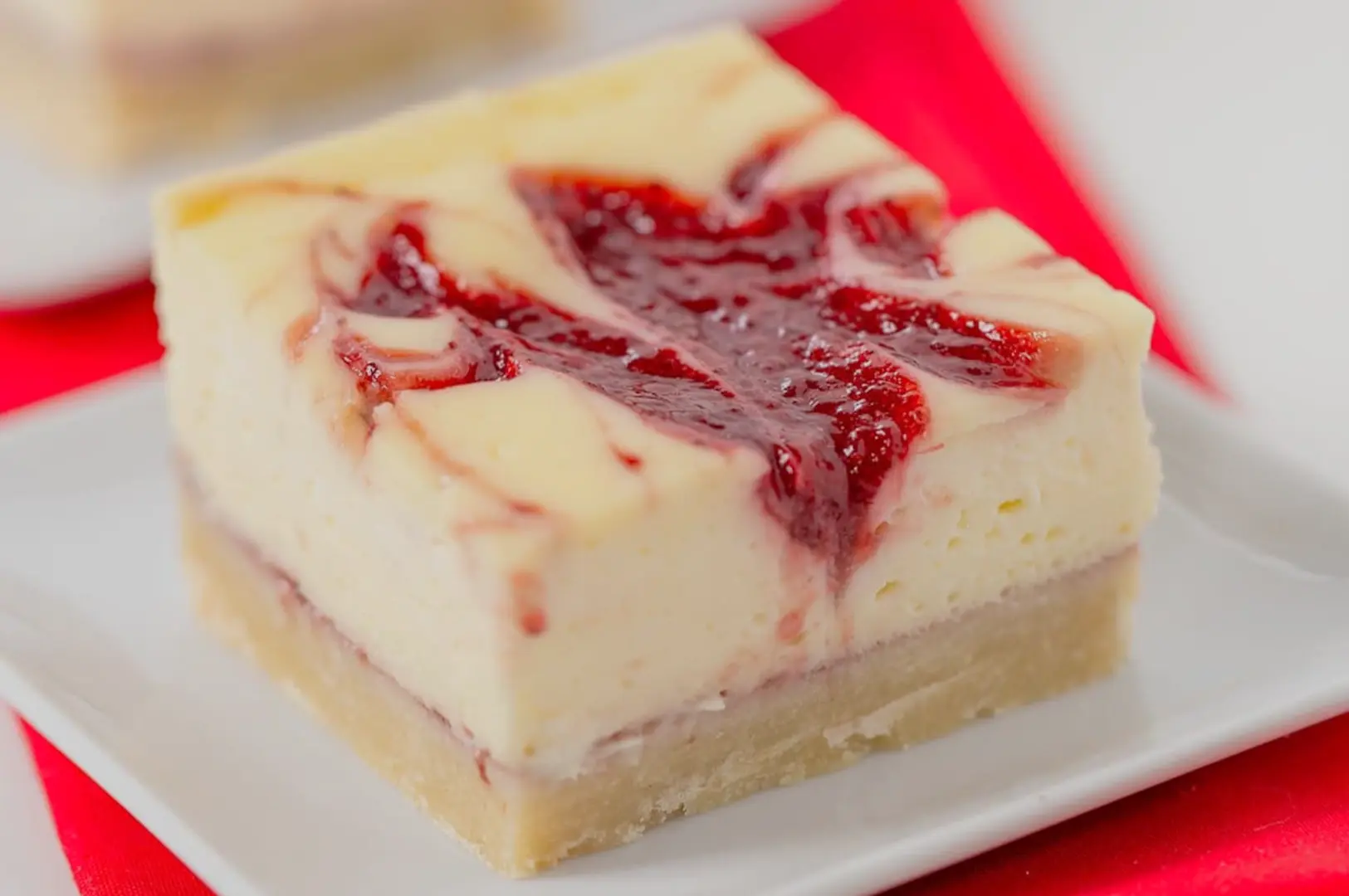 A creamy cheesecake bar with a crumbly golden crust and glossy red berry swirl sits on a white plate against a bright red-and-white backdrop.