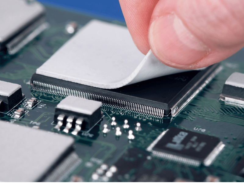 A macro close-up of a human finger peeling back a white thermal pad from a black integrated circuit chip on a green printed circuit board (PCB), showing the intricate solder pins and surface-mount components.