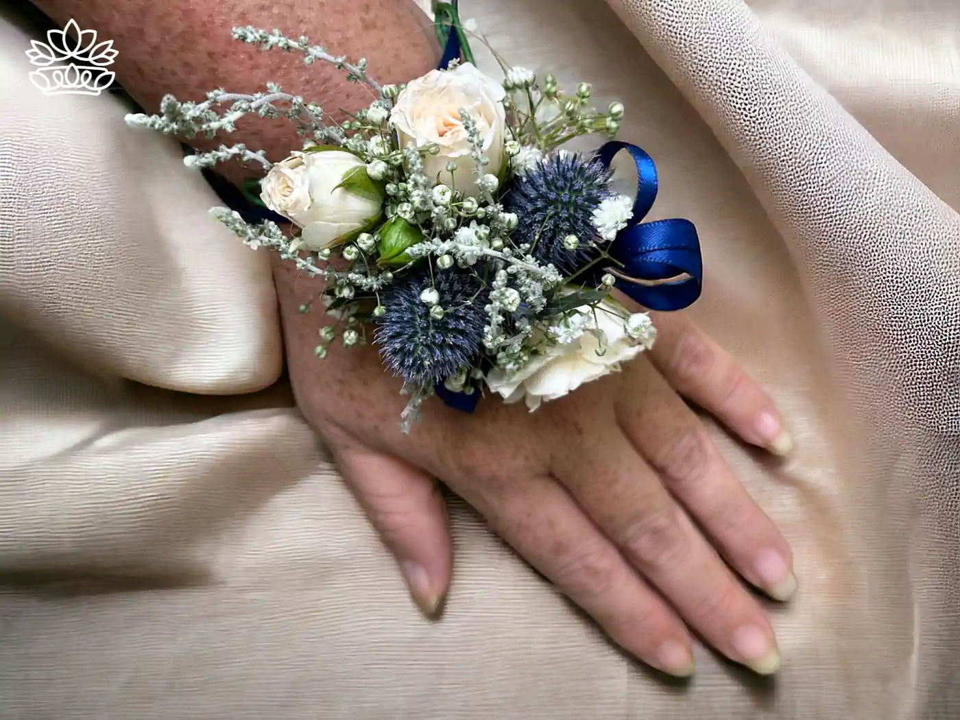 A delicate hand showcases a sophisticated floral corsage featuring ivory roses, striking blue thistle, and soft baby's breath, tied with a deep blue ribbon against a silky beige background. Fabulous Flowers and Gifts - Matric Dance. Delivered with Heart.