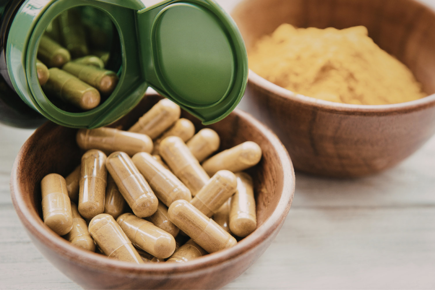 Wooden bowls containing capsules and powder sit on a white surface next to a green bottle.