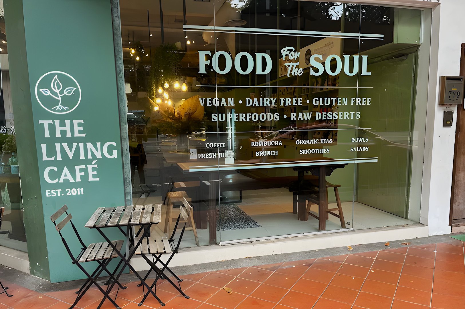 Exterior of The Living Café with a small outdoor table and chairs. The window displays text on vegan and gluten-free options, like superfoods and smoothies.