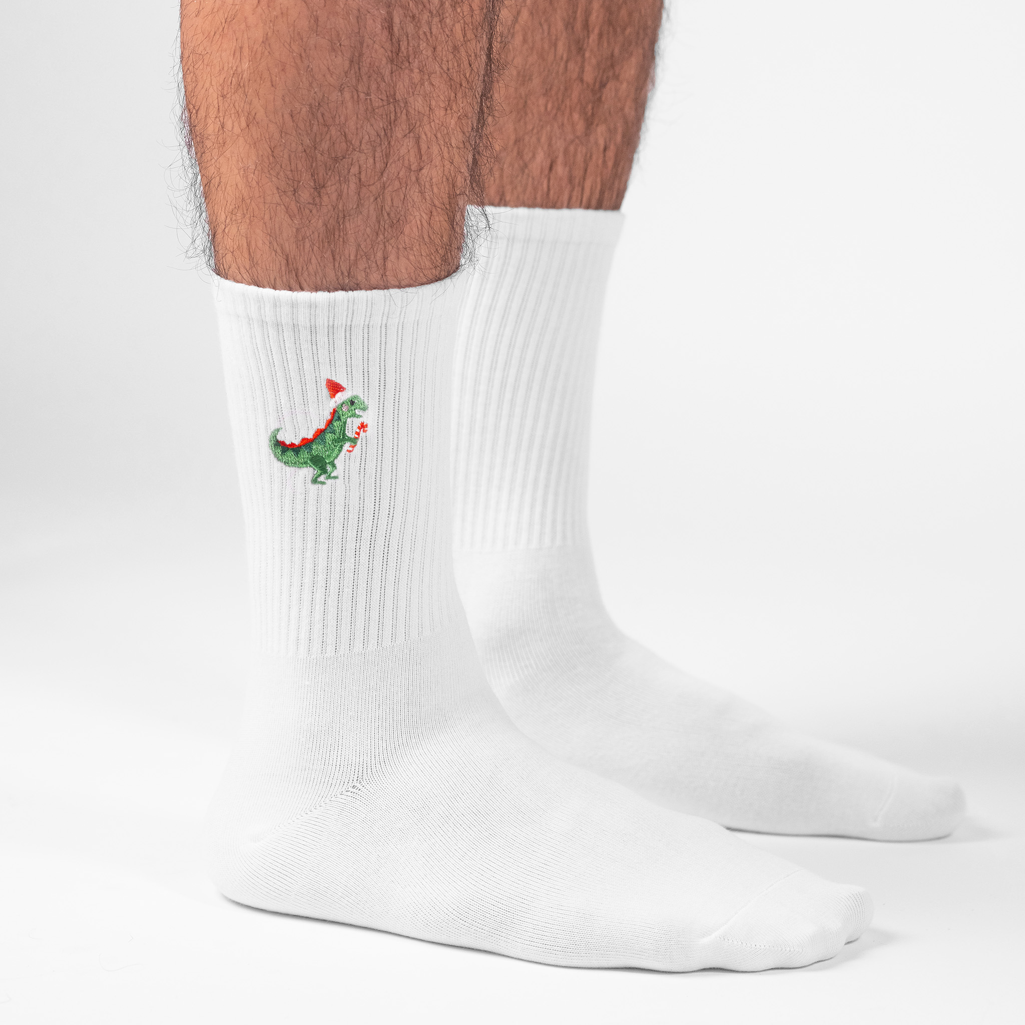 A male model wearing a pair of personalised white crew socks with the an dinosaur wearing a Santa hat and holding a candy cane embroidered onto each sock.