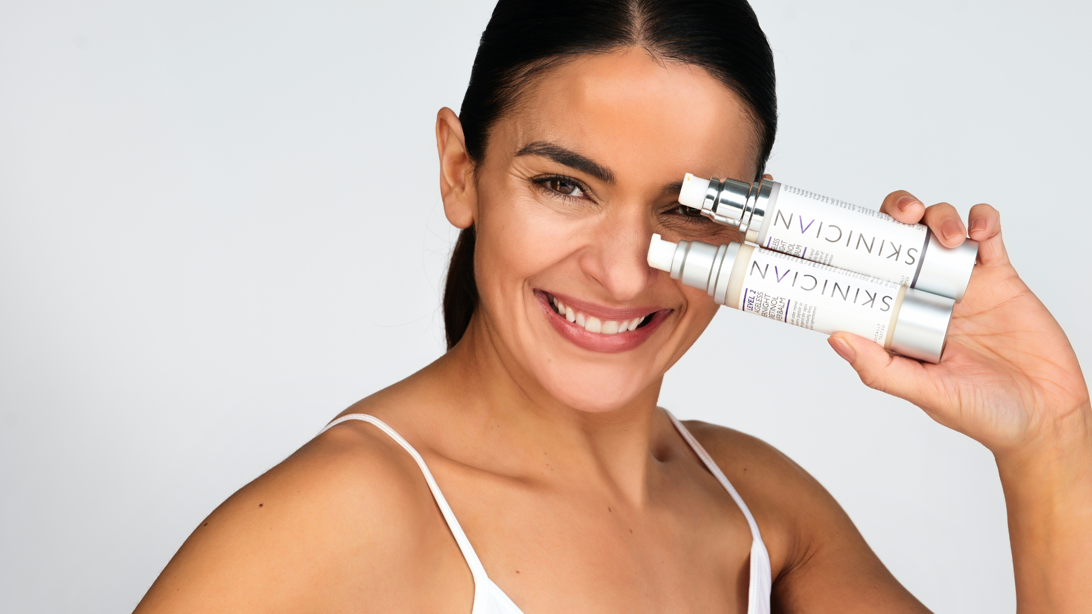 A woman holding SKINICIAN's Overnight Retinol Powerbalm. She is holding the creams to her face and looks happy.