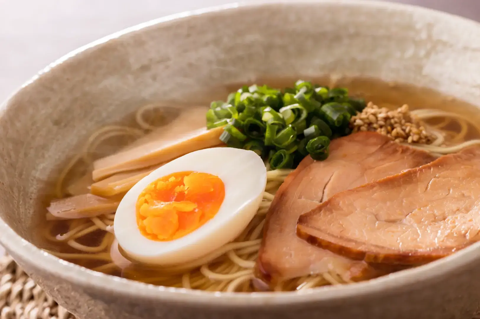 Close-up of ramen bowl with soft-boiled egg, chashu pork slices, scallions, sesame seeds, and thin noodles in clear broth.