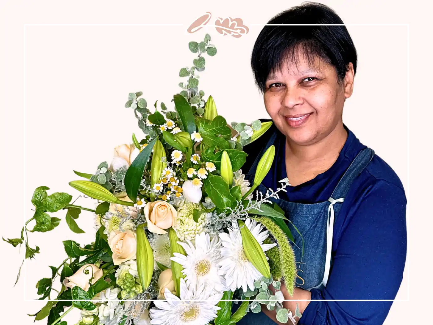 A smiling woman in a blue apron holding a large bouquet of white lilies, roses, and daisies with green foliage, against a white background. Fabulous Flowers and Gifts.