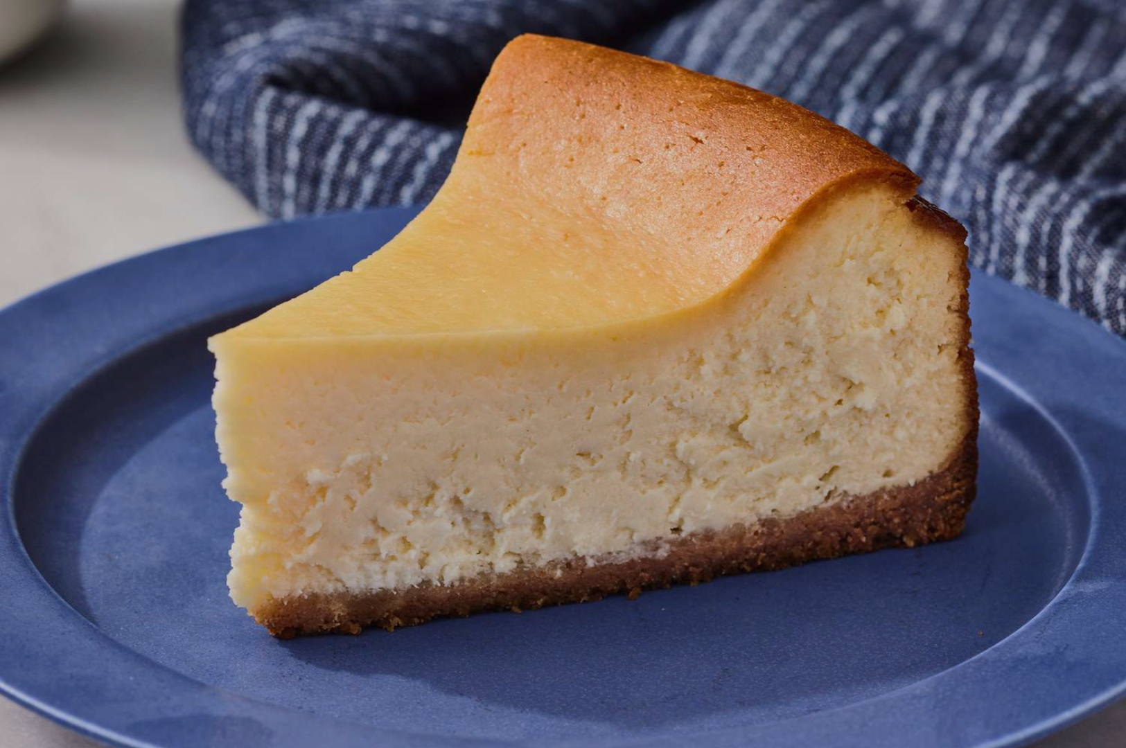Slice of creamy cheesecake with a golden top, resting on a blue plate. A textured, dark blue cloth is in the background, creating a cozy feel.