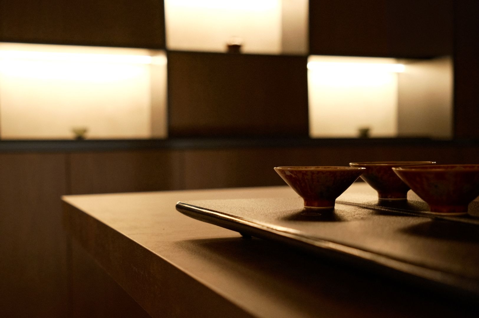 Dimly lit, modern interior with soft, warm lighting highlighting minimalist wooden shelves. A tray with small bowls rests on a sleek wooden table.