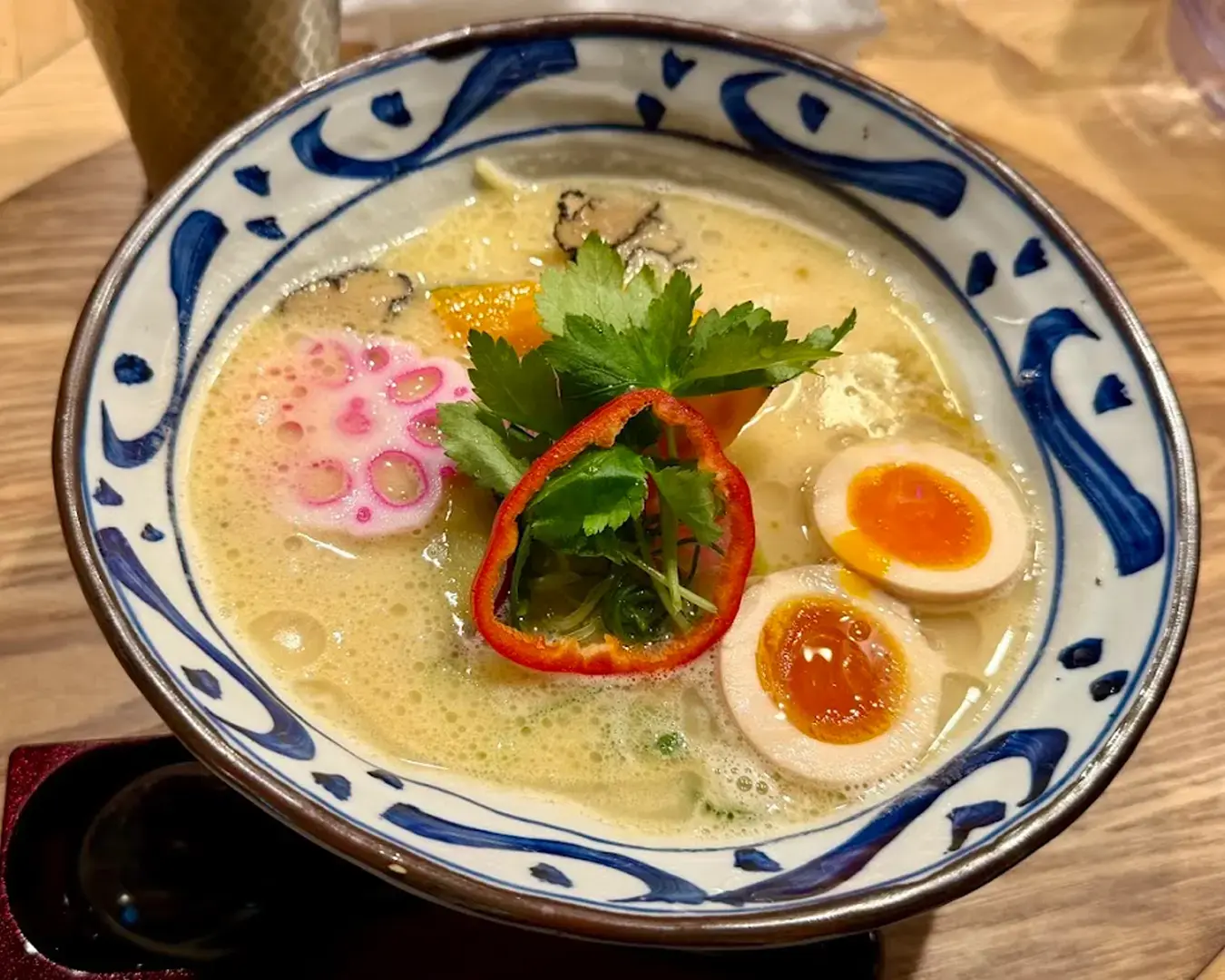 A bowl of creamy ramen with soft-boiled eggs, pink fish cake, cilantro, and sliced red pepper garnished in a blue-patterned bowl on a wooden table.