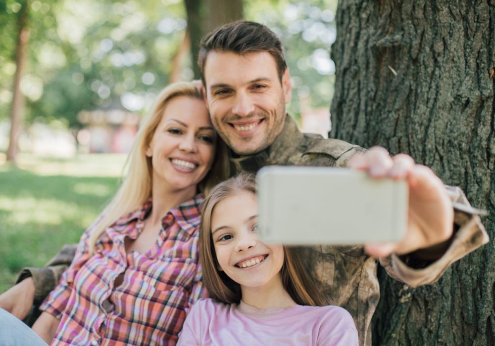 Cute young family of three snapping a selfie leaning against a tree.