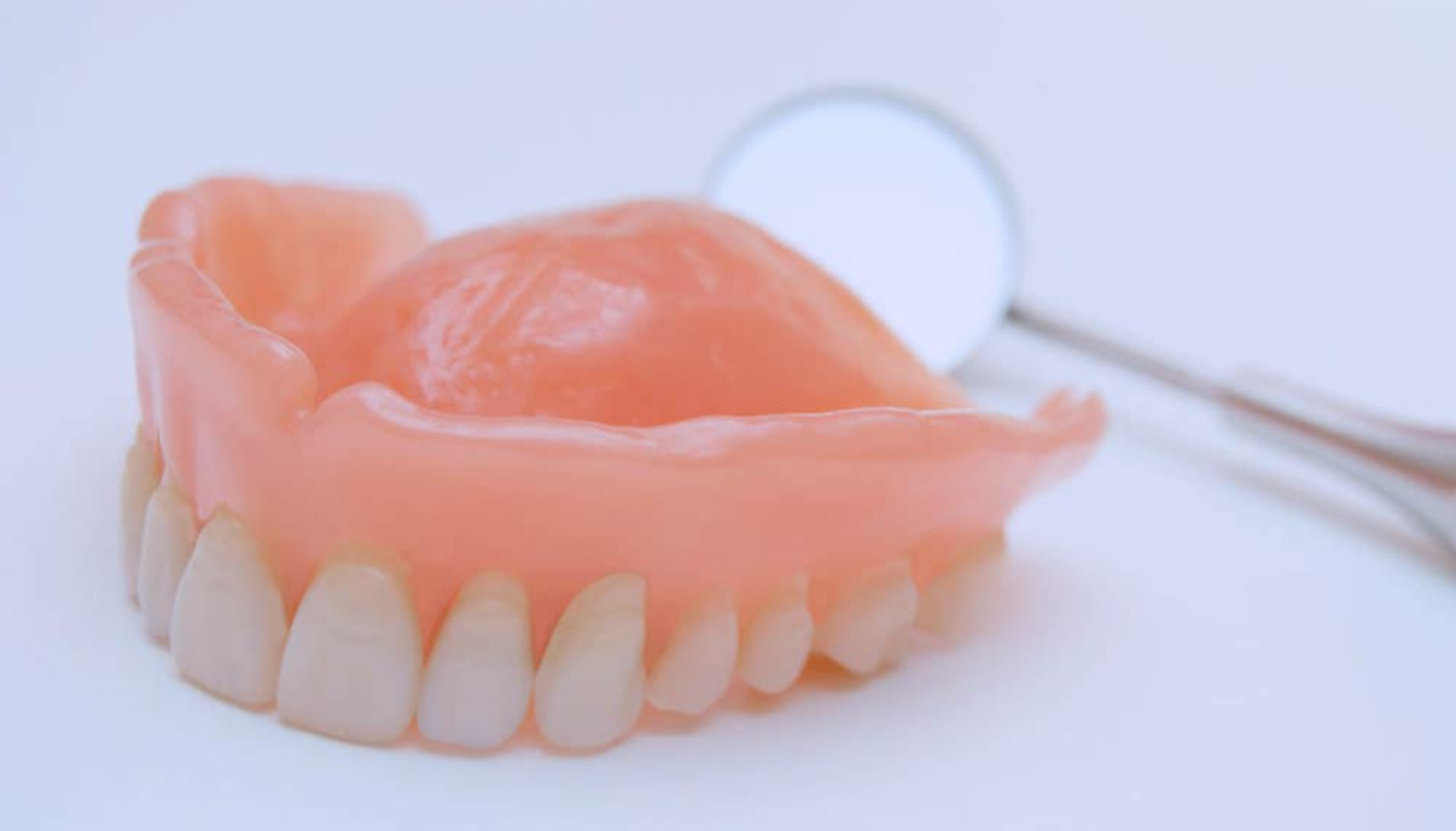 Hel overkjeve tannprotese med rosa tannkjøttbase og kunstige tenner, plassert på hvit overflate ved siden av et tannlegespeil. Ask ChatGPT