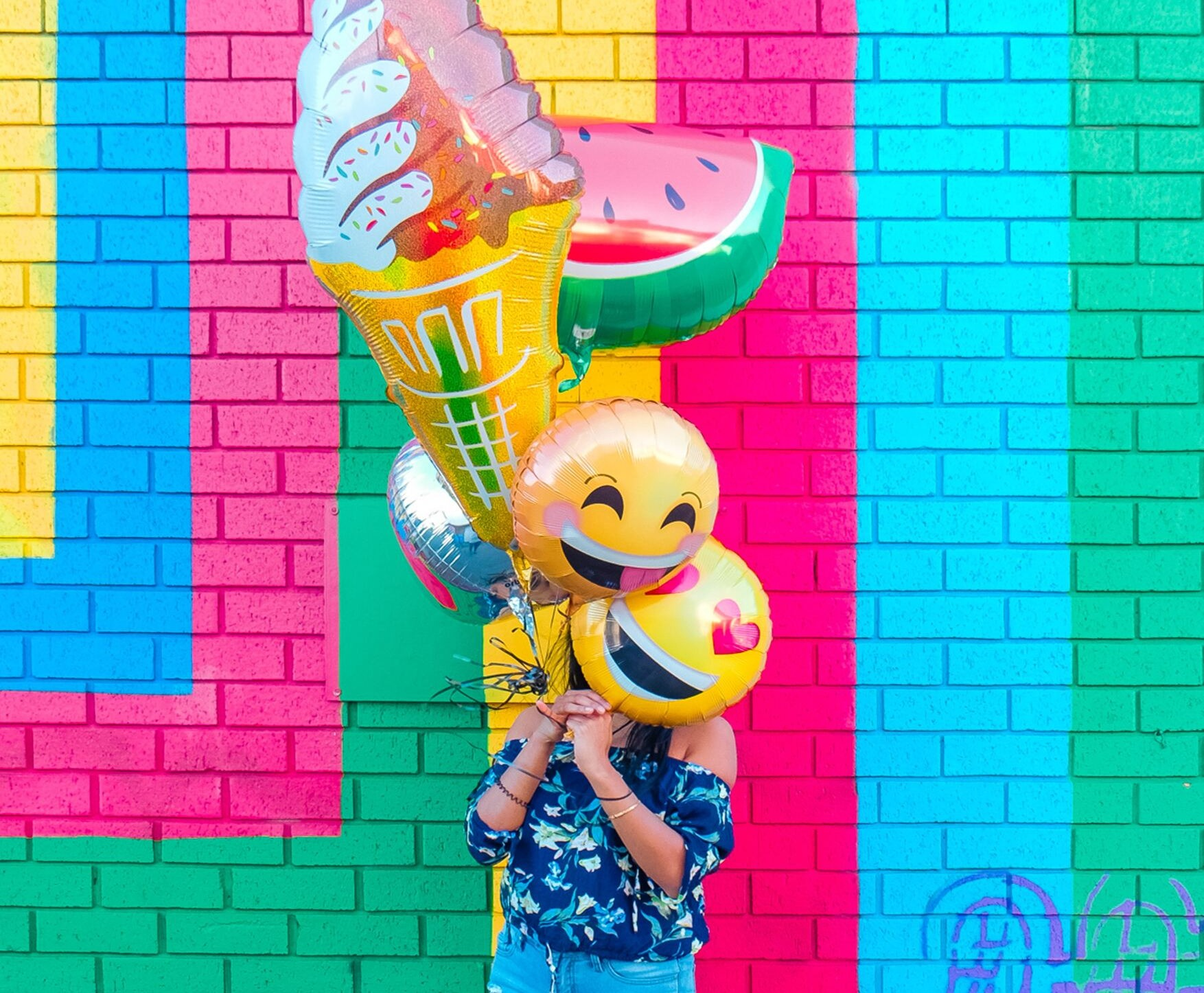 Vrouw staat voor een gekleurde muur en houdt ballonnen voor haar gezicht in de vorm van smiley gezichtjes, een room ijsje en een schijf water meloen.