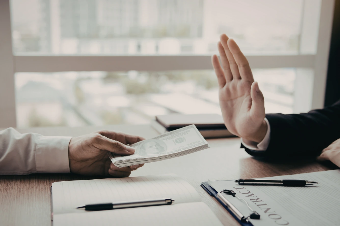 A hand rejects a stack of money, with a contract and pen visible on the desk.