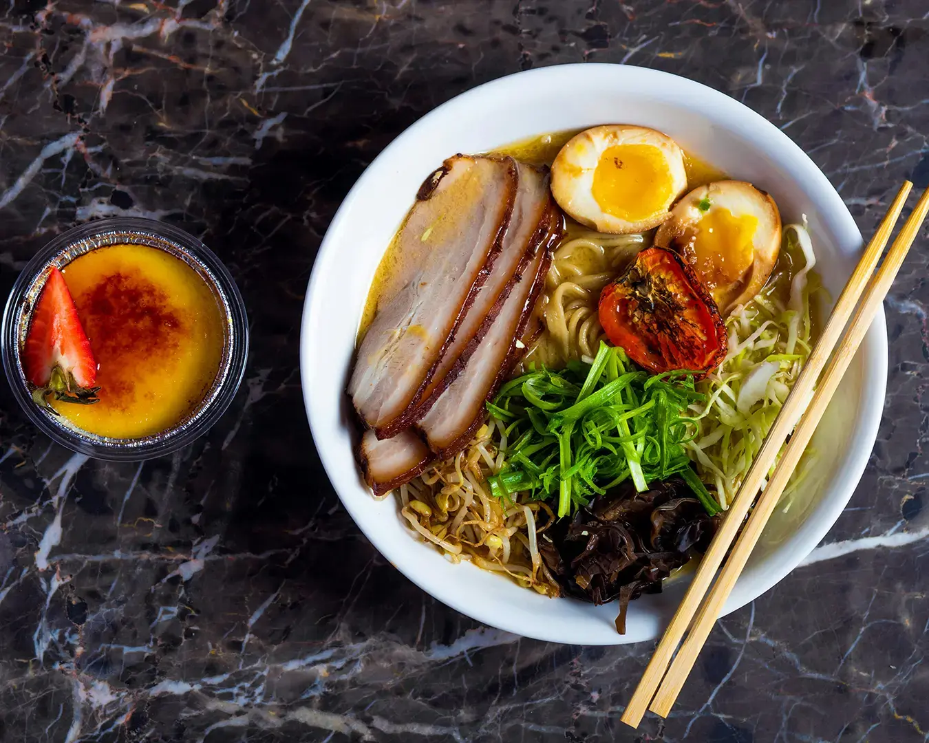 A bowl of ramen with sliced pork, a halved soft-boiled egg, greens, and roasted tomato, alongside crème brûlée with a strawberry on a marble surface.