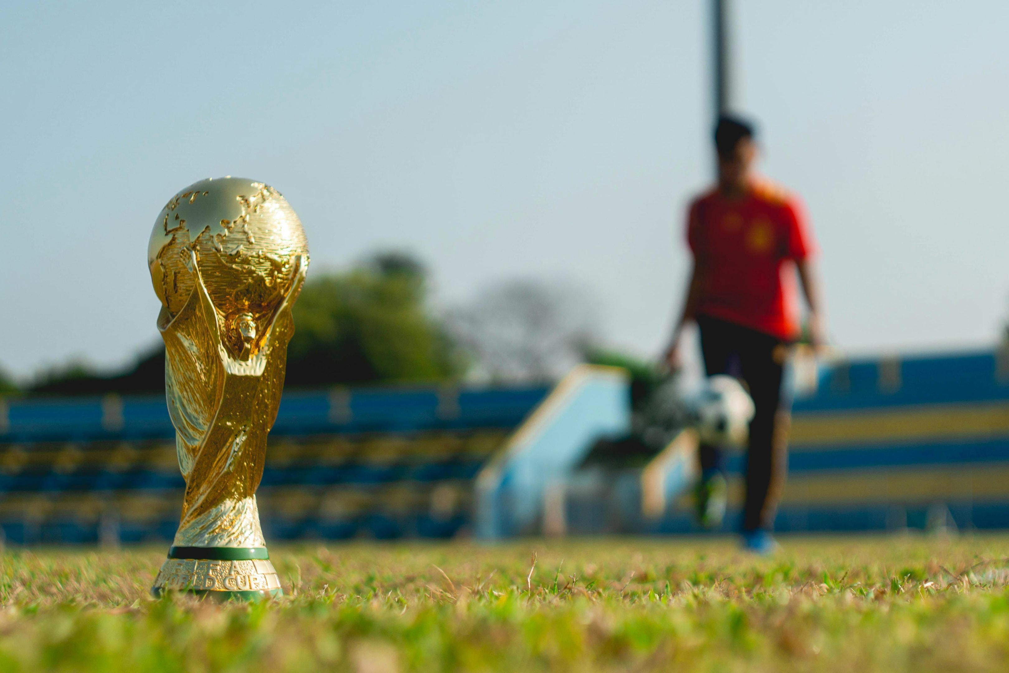 A golden replica of the World Cup trophy stands in sharp focus on the grassy field, gleaming under the bright sunlight. In the blurred background, a figure wearing a red shirt walks across the pitch near a section of blue stadium seating.