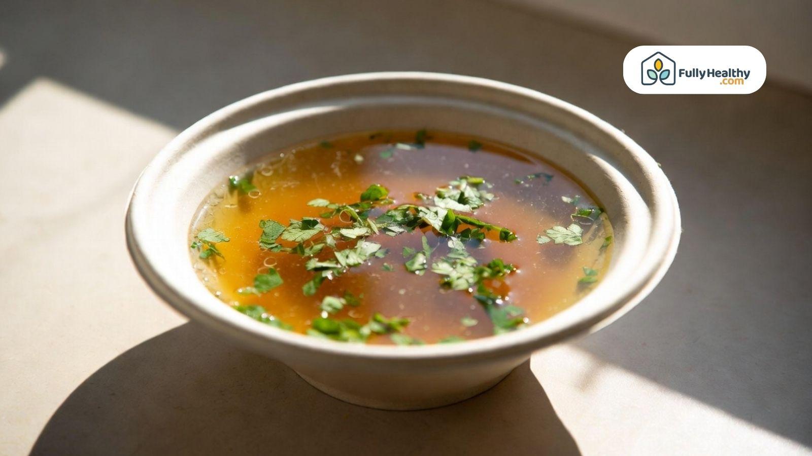 Bowl of clear broth with fresh herbs in natural sunlight.