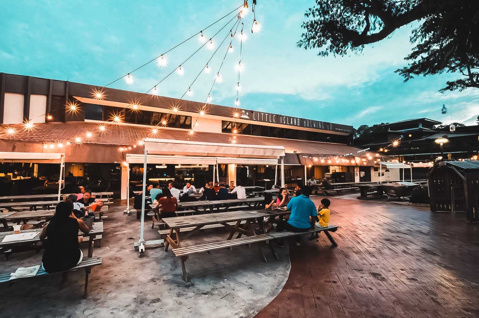 Outdoor dining area at Changi Village featuring string lights, wooden picnic tables, and a lively evening atmosphere.