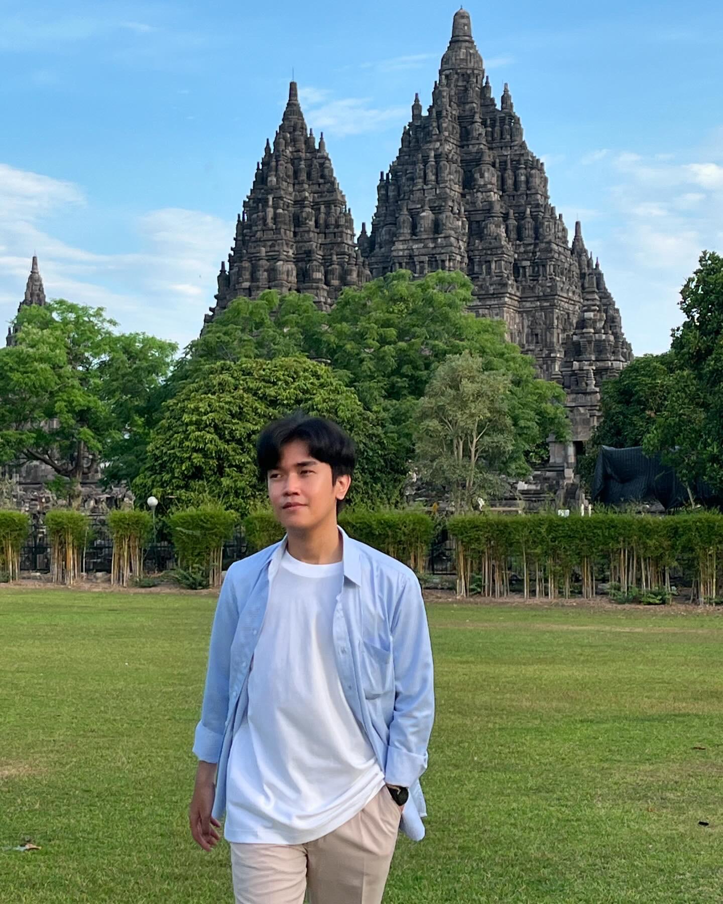 A young man in a light blue open shirt over a white tee stands on the grounds of Prambanan temple complex in Java, Indonesia — the towering 9th-century Hindu stone spires rise dramatically behind him against a partly cloudy sky, evoking a sense of cultural heritage and personal connection to place.