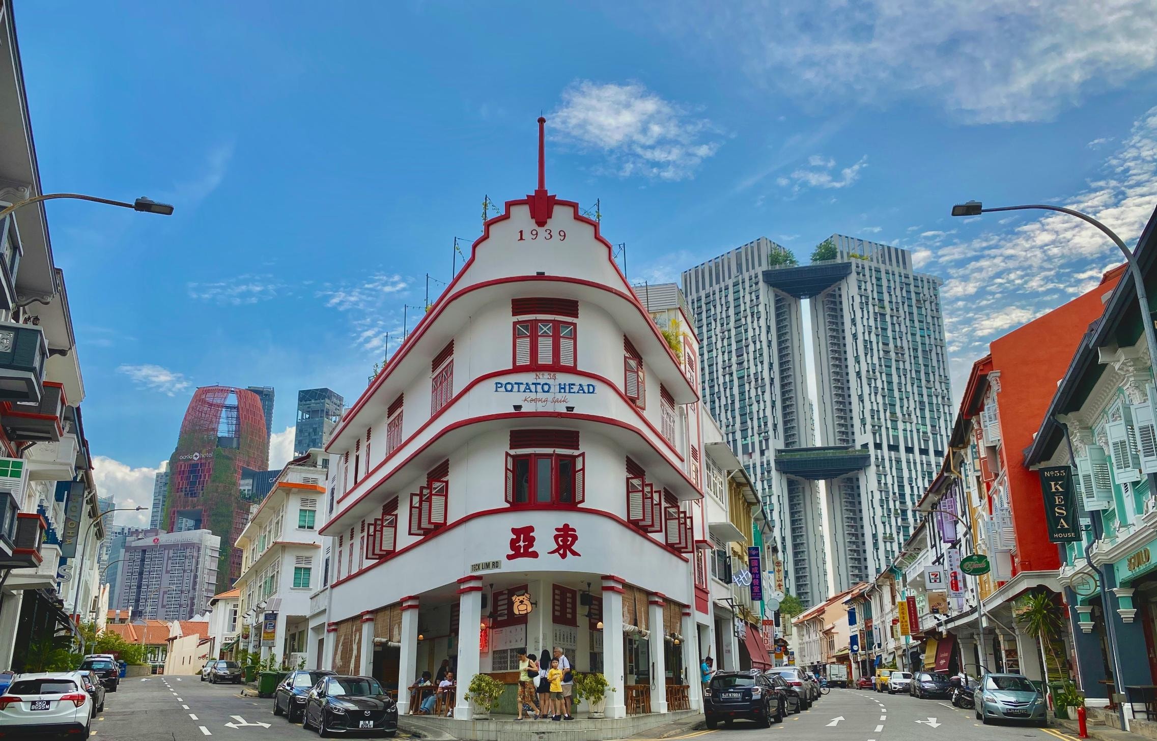 A distinctive white Art Deco building with vibrant red shutters and the year "1939" on its facade stands prominently at a street corner, housing a venue labeled "Potato Head". Behind this historic landmark and the surrounding colorful shophouses, towering modern skyscrapers rise against a bright blue sky.