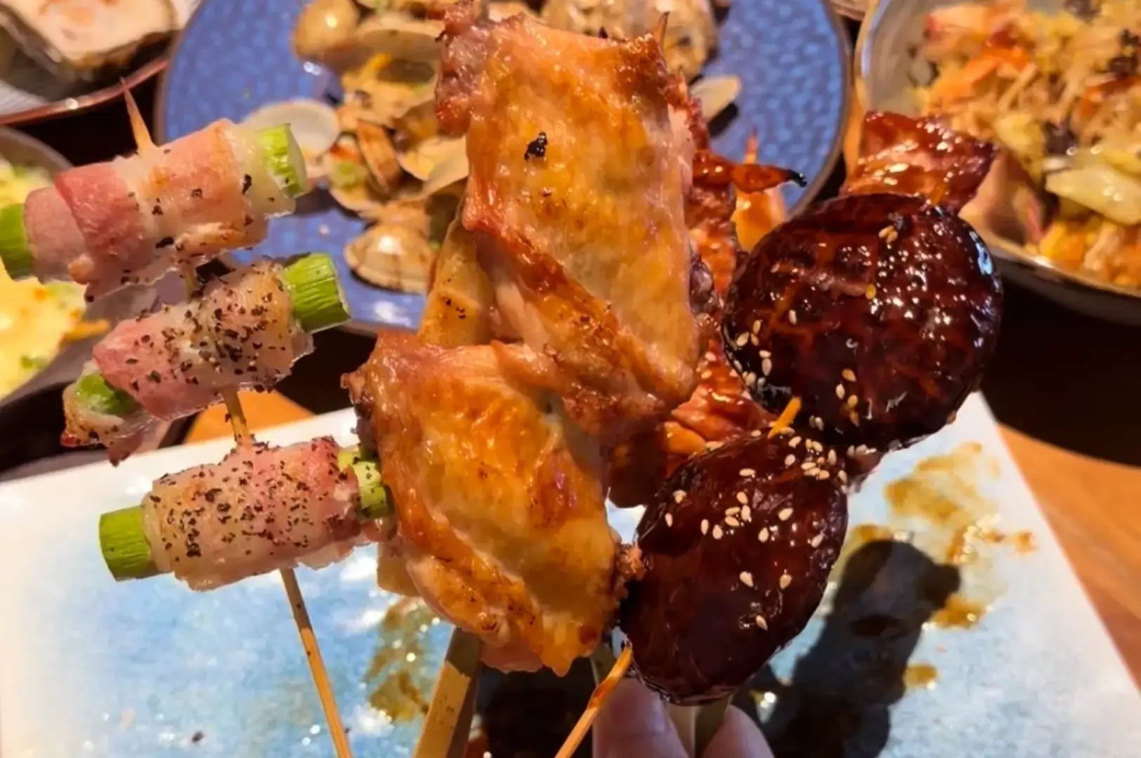 Assorted grilled skewers on a blue plate, including bacon-wrapped vegetables, grilled chicken, and glossy meatballs sprinkled with sesame seeds.