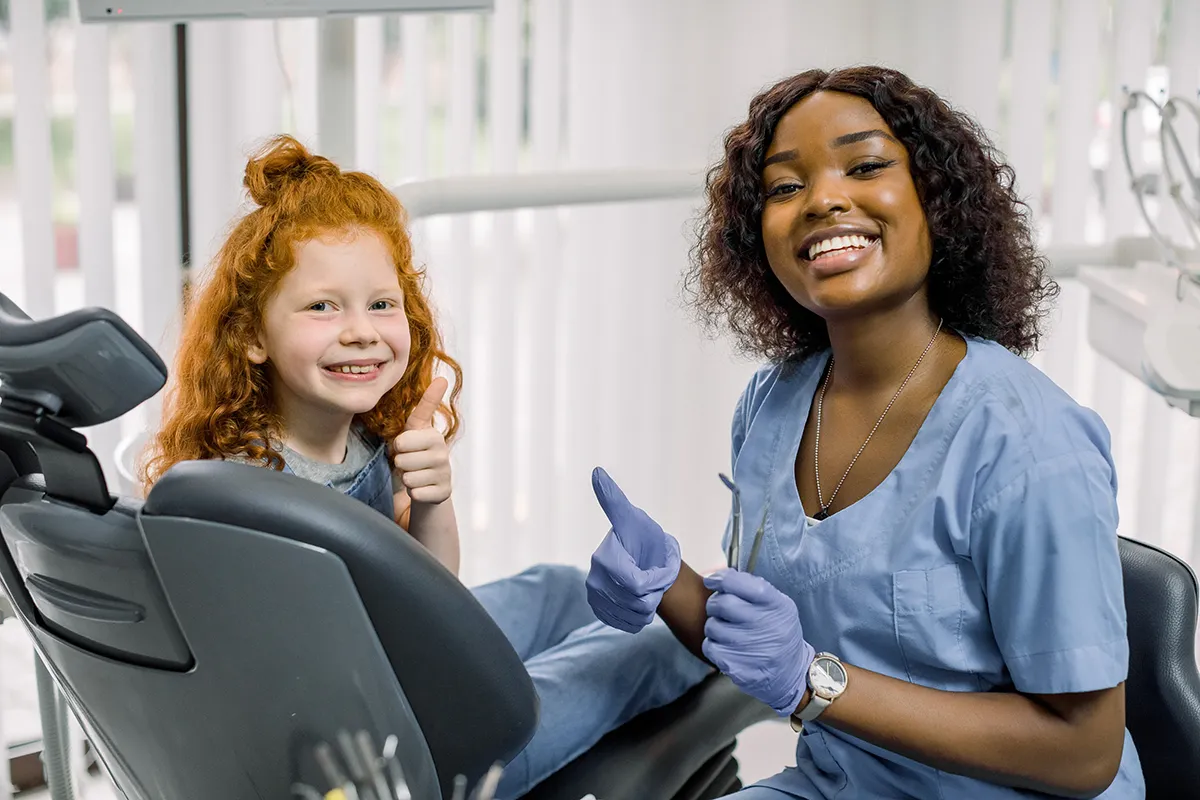 Young child gives thumbs up next to smiling dental professional in bright office.