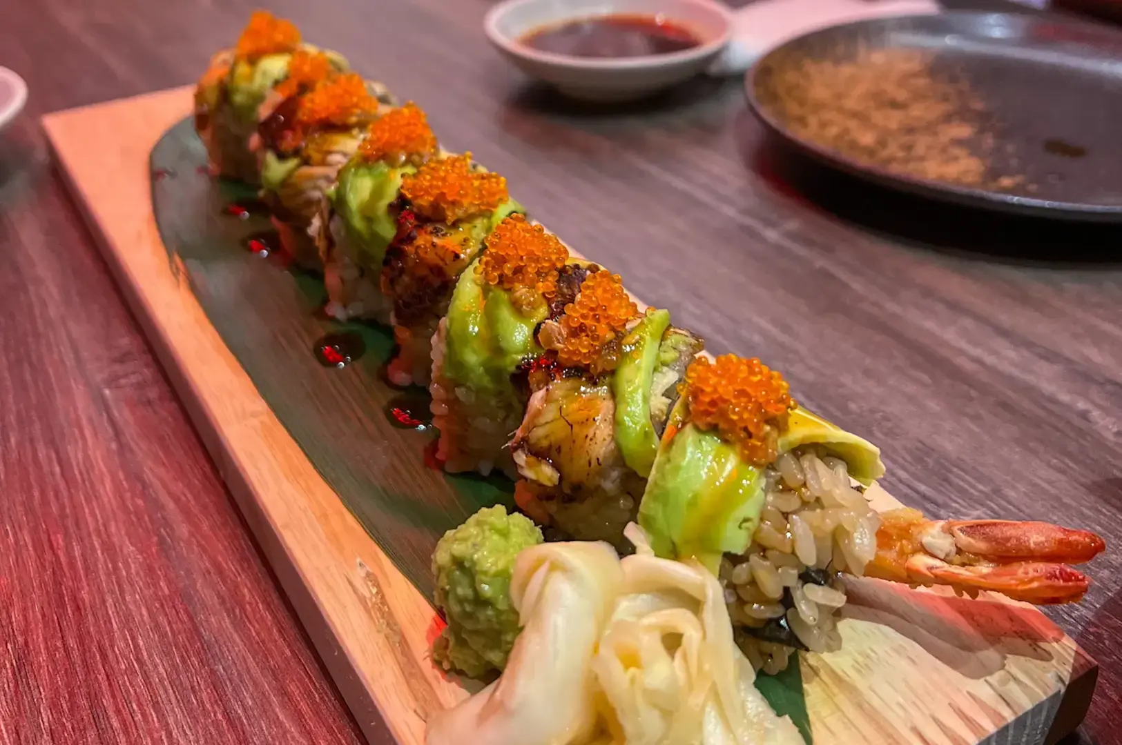 Sushi roll topped with avocado and orange fish roe, arranged on a wooden serving board with pickled ginger and dipping sauce.