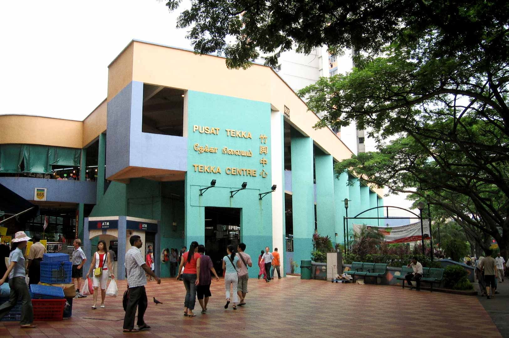 Tekka Centre in Singapore, vibrant multicultural hub with pastel-colored facade and signage in Malay, Tamil, English, and Chinese, surrounded by plaza activity and shaded by large trees.