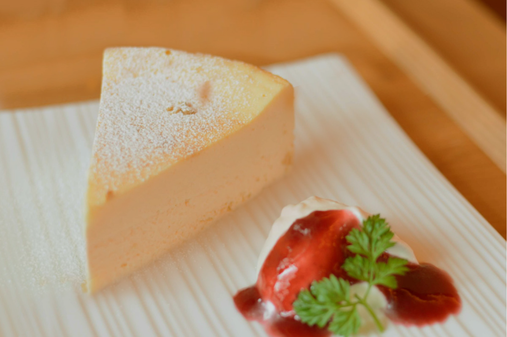 A slice of creamy cheesecake dusted with powdered sugar on a white plate, beside a dollop of whipped cream, strawberry sauce, and a sprig of parsley.