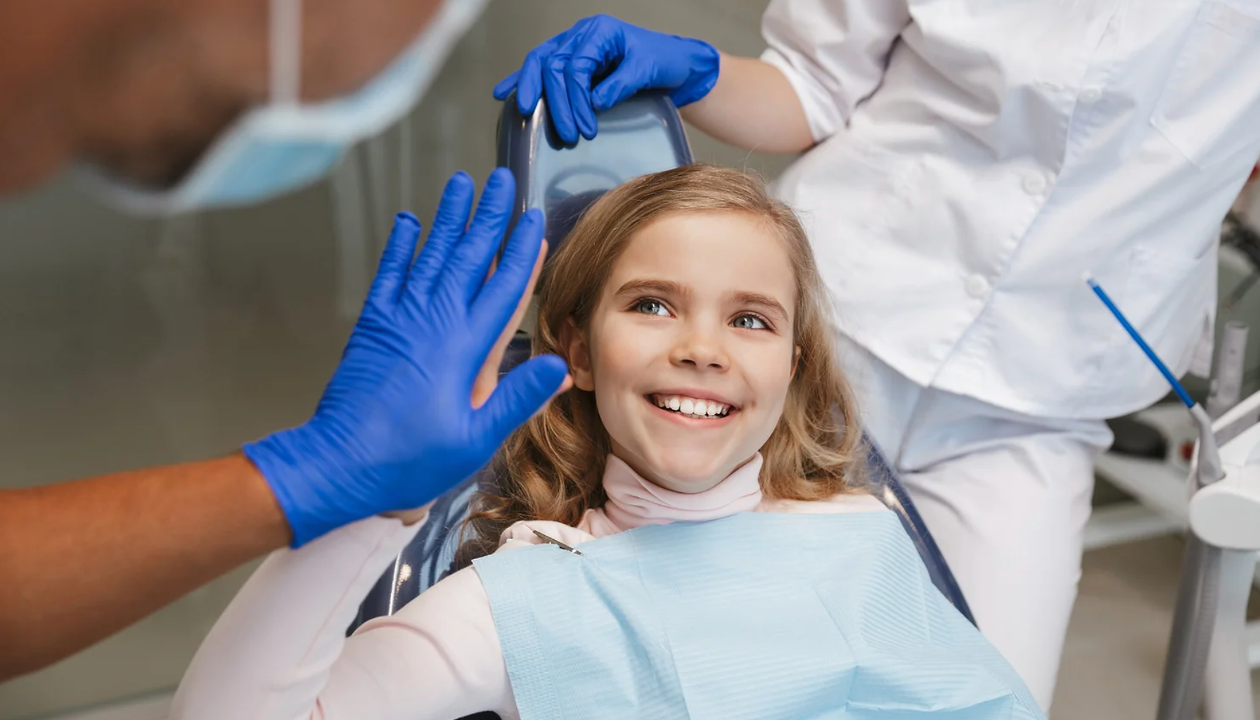 Kul i tannkjøttet – Bør du bekymre deg? Dette må du vite 13 Smilende jente sitter i tannlegestol og gir high five til tannlegen med blå hansker. Tannhelsepersonell i bakgrunnen, vennlig og støttende atmosfære.