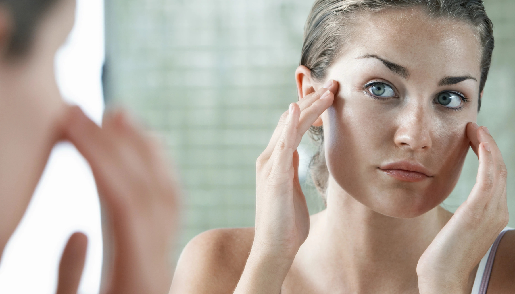 Ung kvinne ser på seg selv i speilet og undersøker huden i ansiktet, muligens som del av hudpleierutine eller bekymring for hudtilstand.