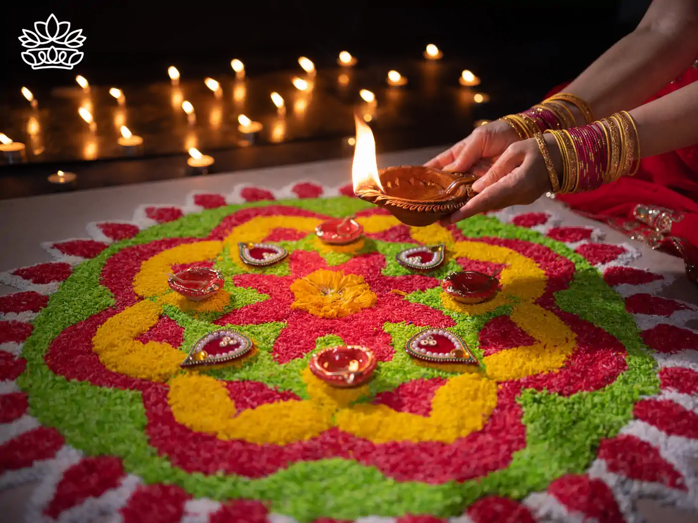 A woman in a red saree carefully placing a lit oil lamp on a beautifully designed rangoli made of vibrant green, red, yellow, and white flower petals, enhancing the festive atmosphere of Diwali. Fabulous Flowers and Gifts - Diwali Flowers. Delivered with Heart.