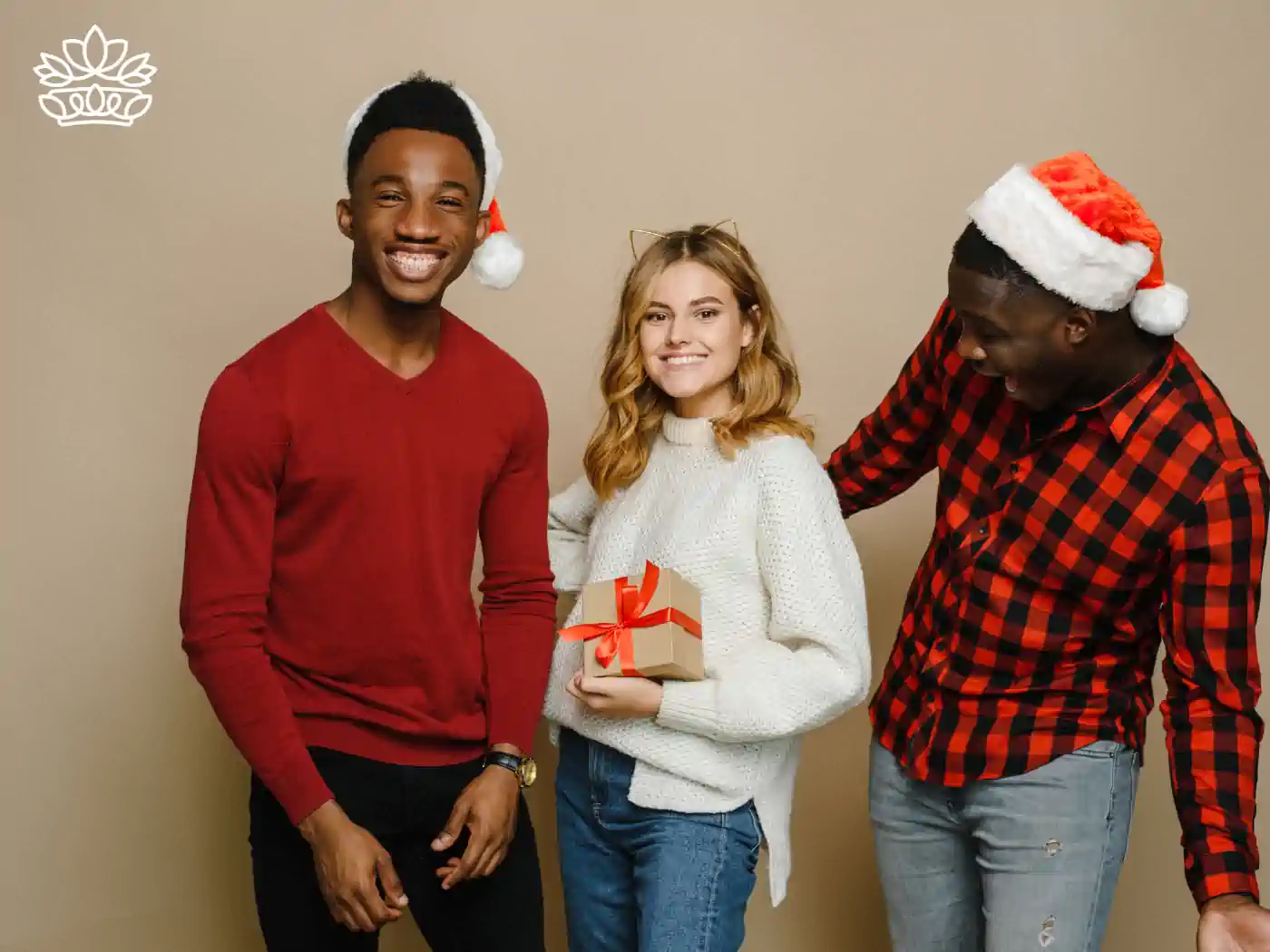 Three friends in festive attire and Santa hats, joyfully posing together, with one holding a gift box wrapped in a red ribbon, part of the Festive Season Gift Boxes Collection. Delivered with Heart by Fabulous Flowers and Gifts.