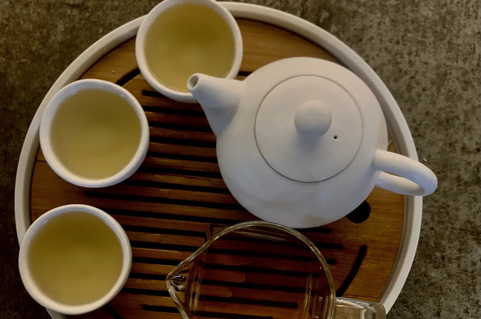 A high-angle, top-down photograph displays a traditional tea service arranged on a circular wooden tray with a slatted bamboo surface. A matte white ceramic teapot with a rounded body and a matching lid sits on the right side of the tray. To the left, three small, handleless white ceramic cups are filled with a pale, golden-green translucent tea. At the bottom of the frame, a clear glass fairness pitcher, or Cha Hai, sits partially on the tray, reflecting the warm, ambient light. The entire tea set rests on a textured, neutral gray stone or concrete countertop, capturing a peaceful and minimalistic tea ceremony aesthetic.