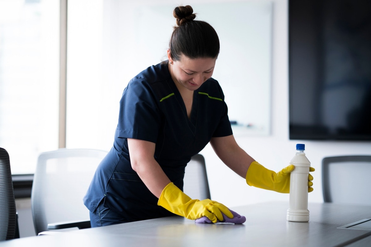 A professional cleaner wearing yellow gloves and a navy uniform wipes down a conference table in a bright, modern office. A bottle of cleaning solution sits nearby