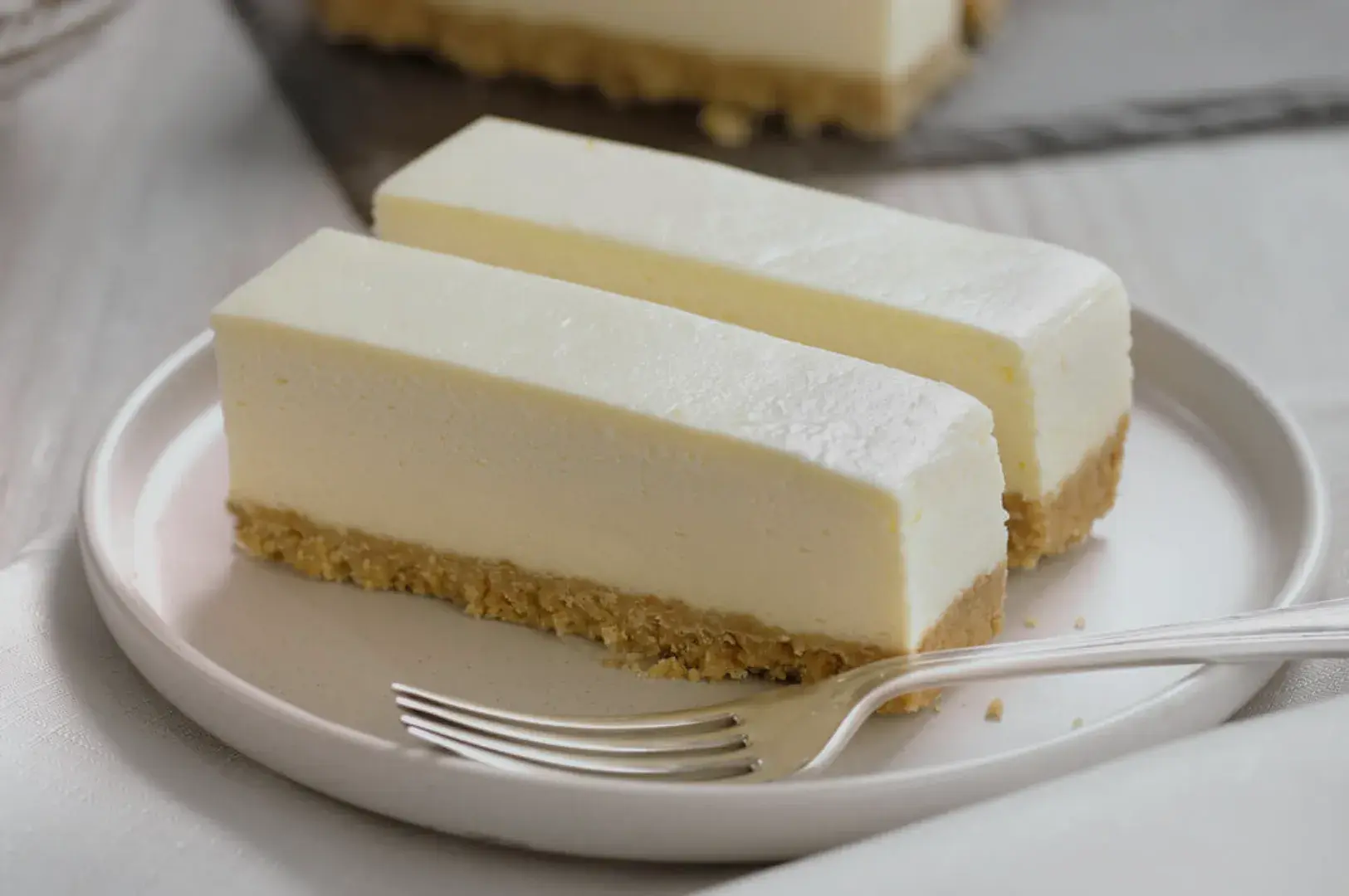 Two rectangular slices of cheesecake on a white plate, with a graham cracker crust and a smooth, creamy top. A fork is placed beside the plate.