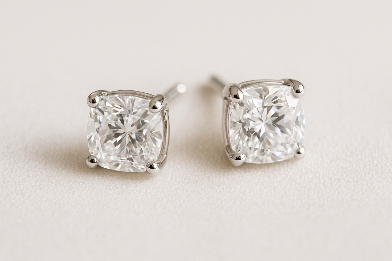 Auskarai su pagalvėlės formos (cushion cut) deimantais, balto aukso įtvirtinimas, blizgantys brangakmeniai ant šviesaus audinio fono.