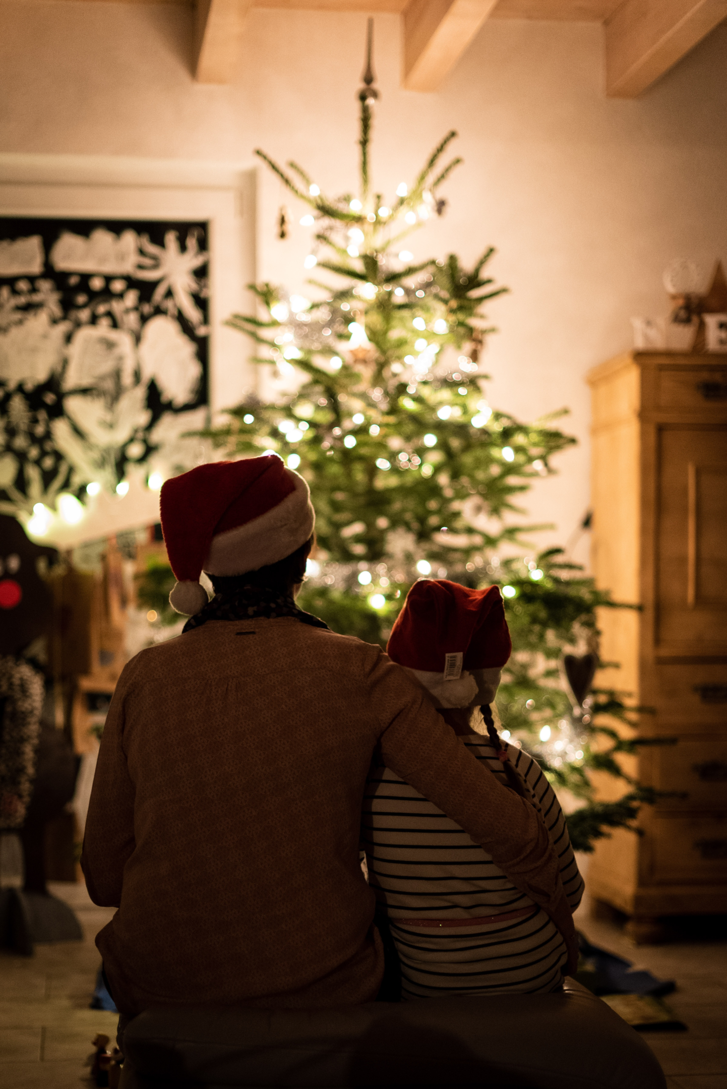 A parent and child facing a lit Christmas tree with their backs to us.