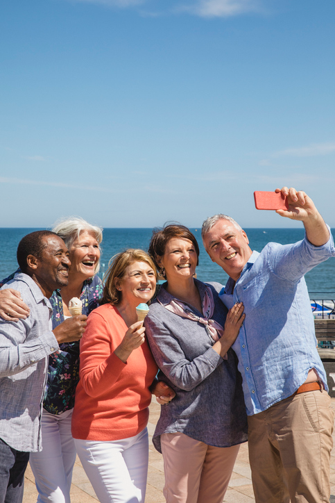 Five mature adults, two eating ice cream cones, snapping a selfie on the beach.