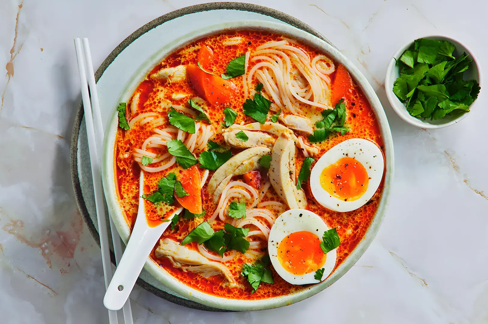 A vibrant bowl of spicy noodle soup with sliced chicken, half-boiled eggs, and fresh cilantro on a marble table. A small dish of herbs is beside it.
