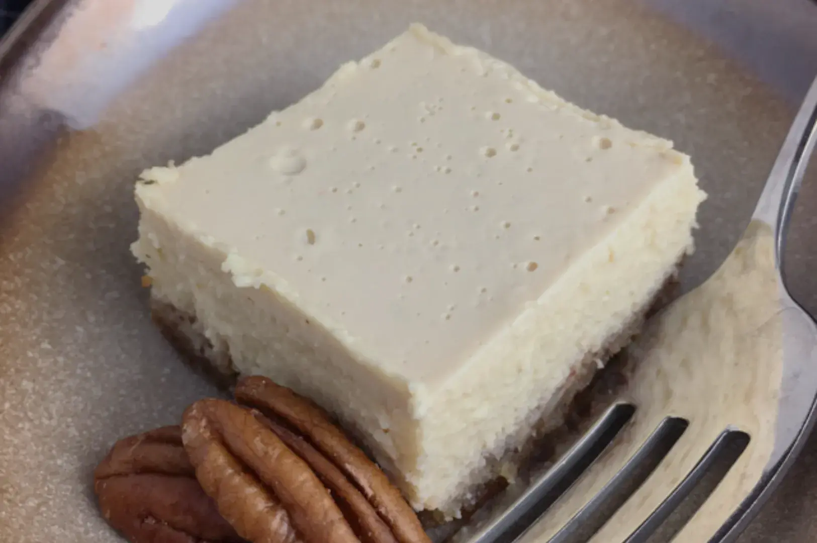 A creamy cheesecake slice with a smooth top and dense texture, garnished with pecans on the side. A fork is placed beside it on a gray plate.
