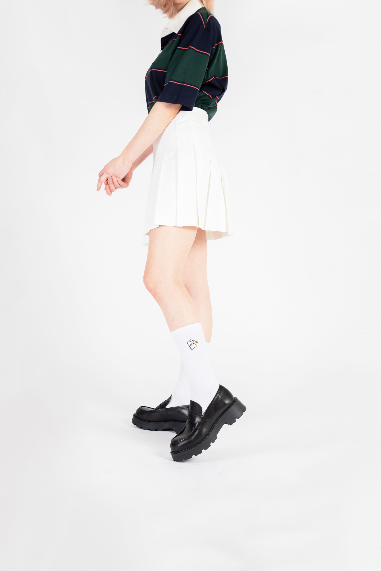 A female model wears a white tennis skirt and rugby polo with black leather loafers and a pair of white crew socks with a a kawaii tea bag embroidered on them..