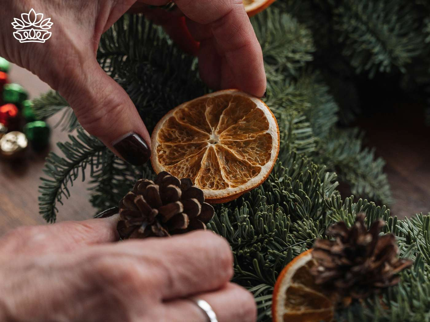Hands meticulously crafting a Christmas arrangement, using hot glue to affix a dried orange slice to a wreath, amidst pine cones and fresh greenery, a festive bow adding the finishing touch, perfect for ideas to hang and enjoy with kids, from the Christmas Wreaths and Flowers Collection at Fabulous Flowers and Gifts.
