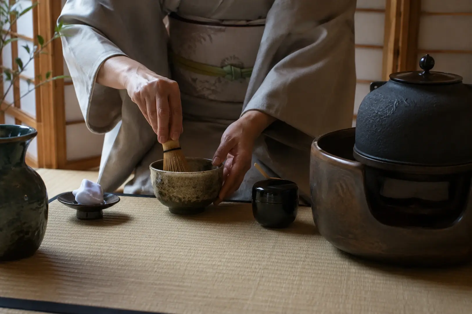 Shows a close-up of a person in a kimono whisking green tea in a mottled ceramic bowl. The scene is laid out on tatami mats with traditional equipment, including a dark iron kettle on a heater, a black lacquer tea container, and a small dish holding a white cloth. The composition emphasizes the delicate hand movements and the focused, ritualistic nature of the preparation.