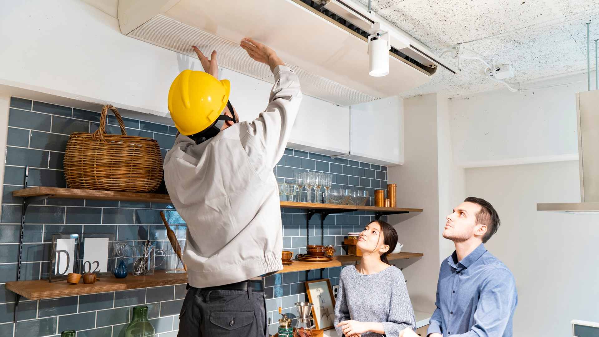 Maintenance technician wearing a yellow hard hat inspecting and servicing a ceiling-mounted HVAC unit while two tenants watch, illustrating professional preventative maintenance for a rental property.
