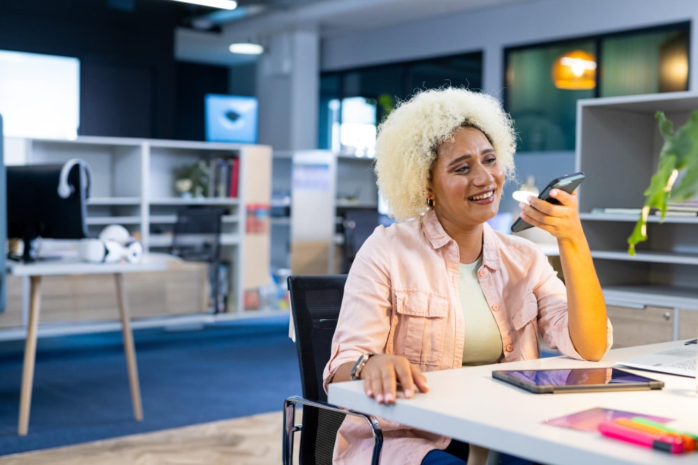 Young professional woman with curly blonde hair using a smartphone and smiling while seated at a desk with digital devices, highlighting flexible business communication through modern phone system upgrades.