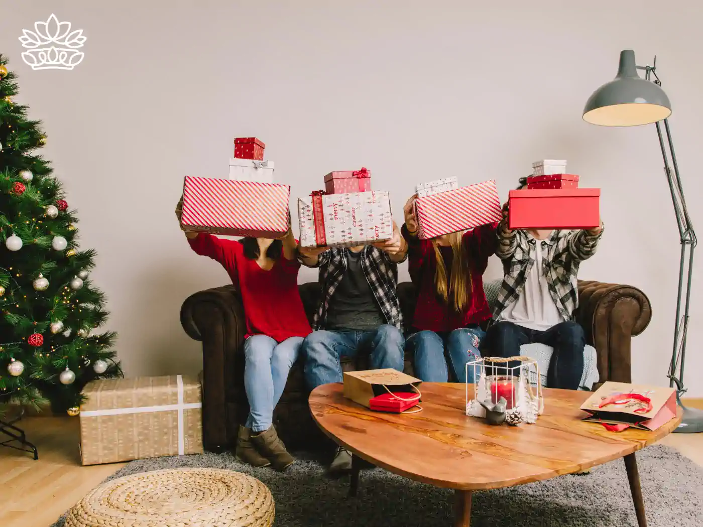 Four friends sitting on a couch, playfully holding up gift boxes in front of their faces, with a Christmas tree nearby, part of the Festive Season Gift Boxes Collection. Delivered with Heart by Fabulous Flowers and Gifts.