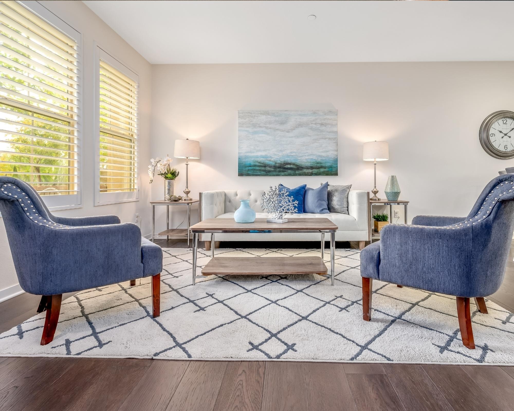 Bright and modern living room with navy blue accent chairs, a white sofa with decorative pillows, a wooden coffee table, and a patterned area rug.