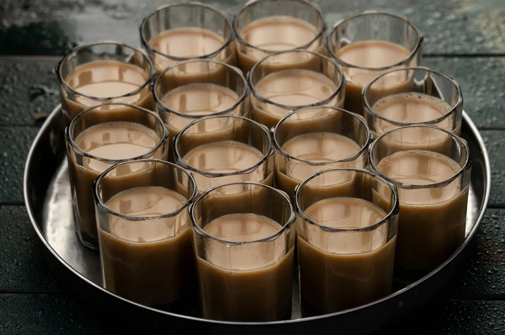 High-angle shot of multiple small, faceted clear glasses filled with warm milk tea or chai arranged neatly on a round metal tray.