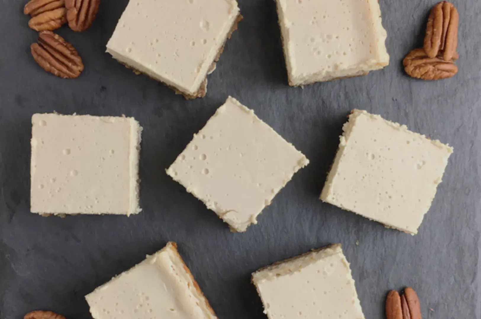 Aerial view of square pieces of cheesecake arranged on a dark surface, with some pecans scattered around.