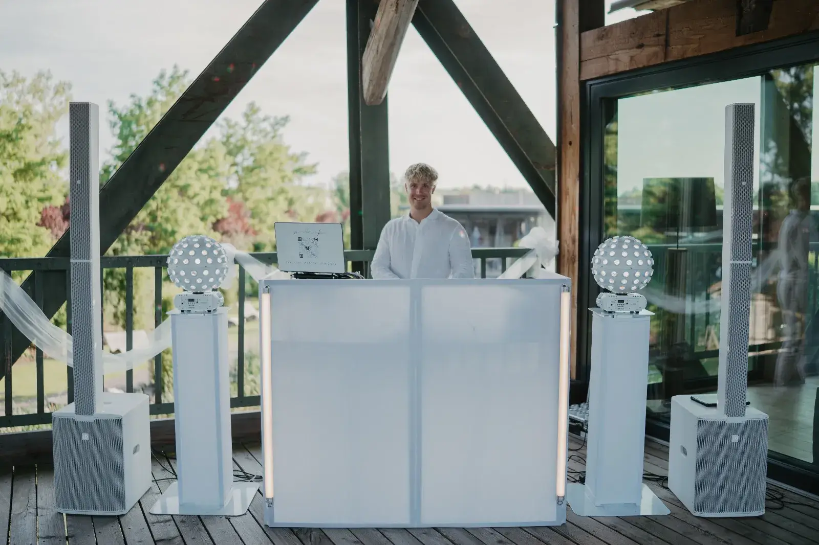 DJ Nathan White in weißem Outfit steht hinter einem modernen weißen DJ-Pult auf einer Terrasse mit Holzträgern; links und rechts befinden sich Lautsprecher und Lichteffekte, im Hintergrund ist eine grüne Außenanlage zu sehen.